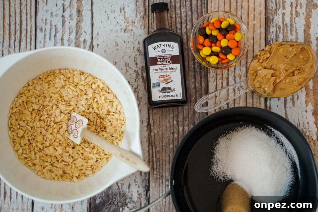 No-Bake Rice Krispies Cookies Recipe 4 A bowl of Rice Krispies, a bottle of vanilla extract, a serving of Reese's pieces, 3/4 cup of peanut butter, and a small saucepan of corn syrup, brown sugar, and granulated sugar.