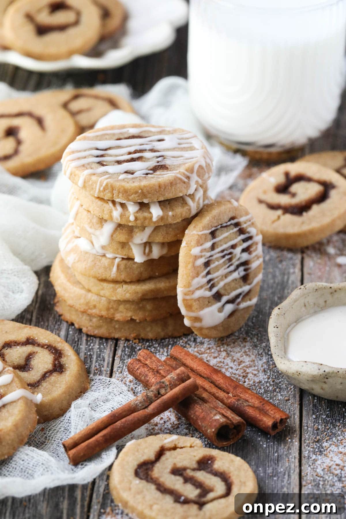 cinnamon roll cookies stacked on a wooden surface with cinnamon sticks and a glass of milk in the corner.