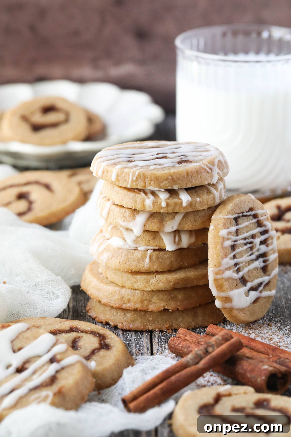 cinnamon roll cookies stacked on a wooden surface with cinnamon sticks and a glass of milk in the corner.