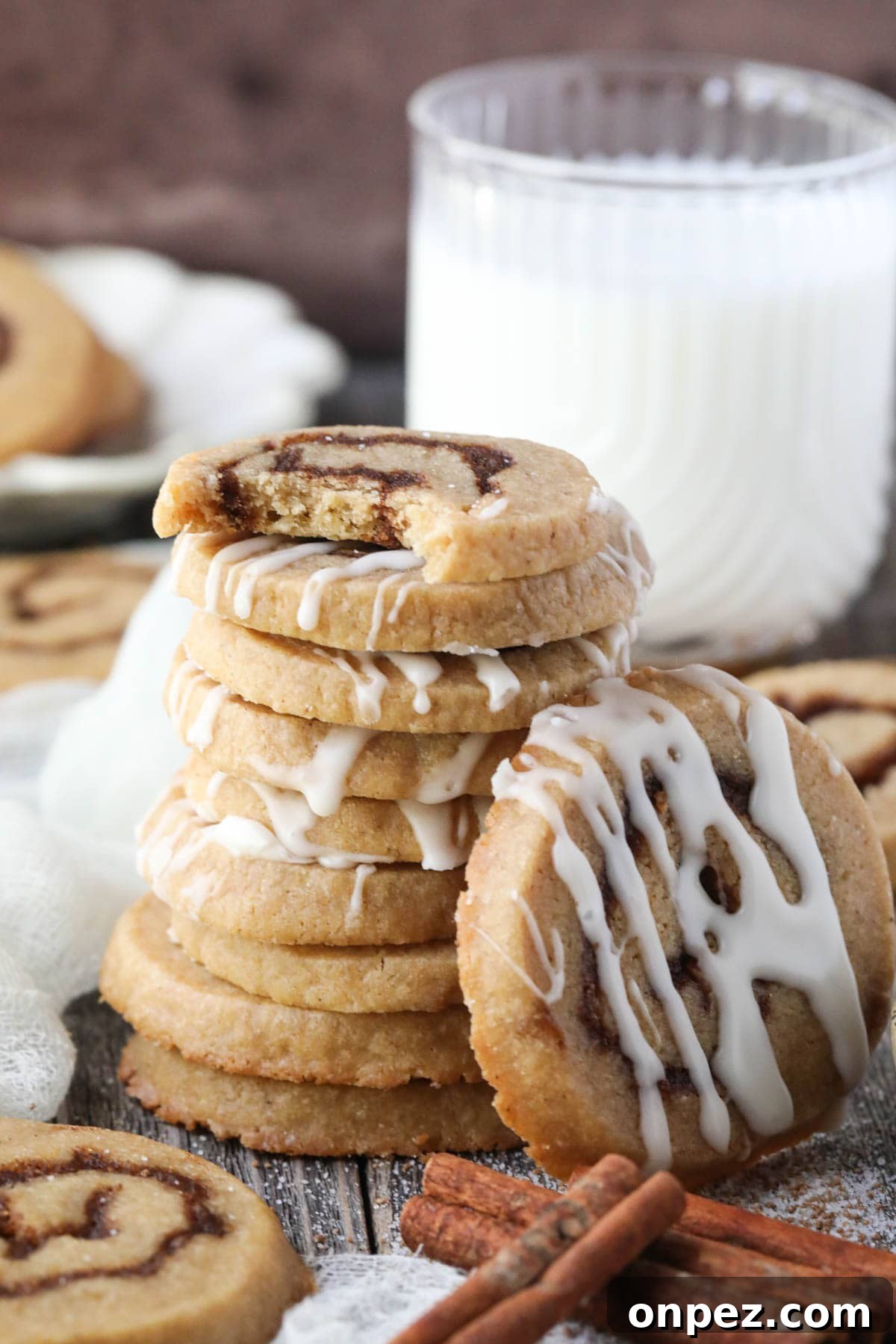 cinnamon roll cookies stacked on a wooden surface with cinnamon sticks and a glass of milk in the corner.