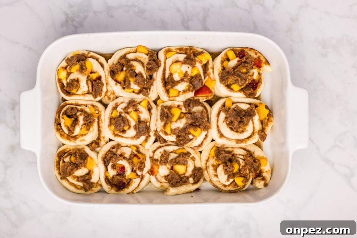 Unbaked cinnamon rolls with peaches and crumble in a white dish.