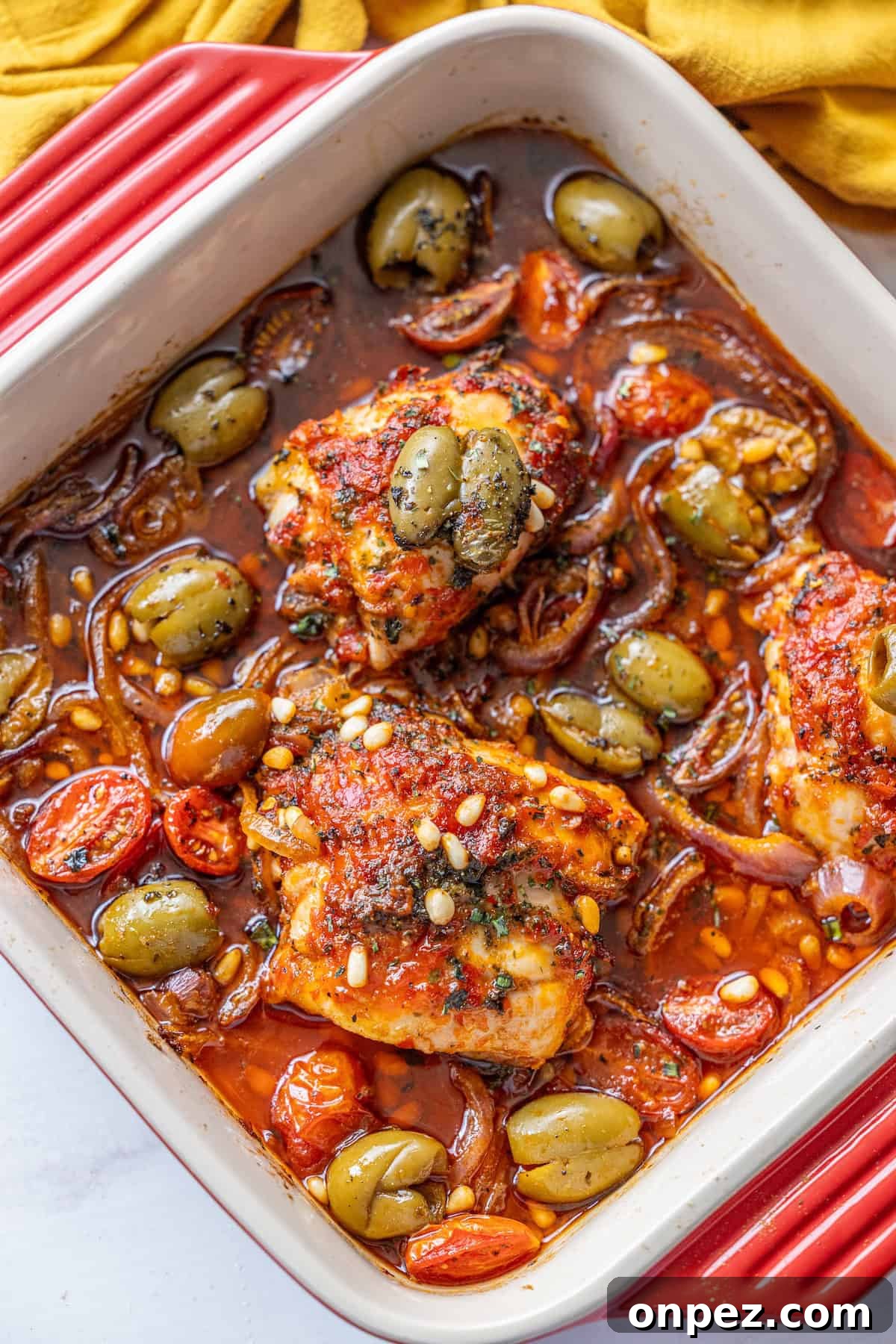 Baked chicken thighs in tomato sauce with green olives, cherry tomatoes, red onions, and pine nuts in a rectangular red and white baking dish.