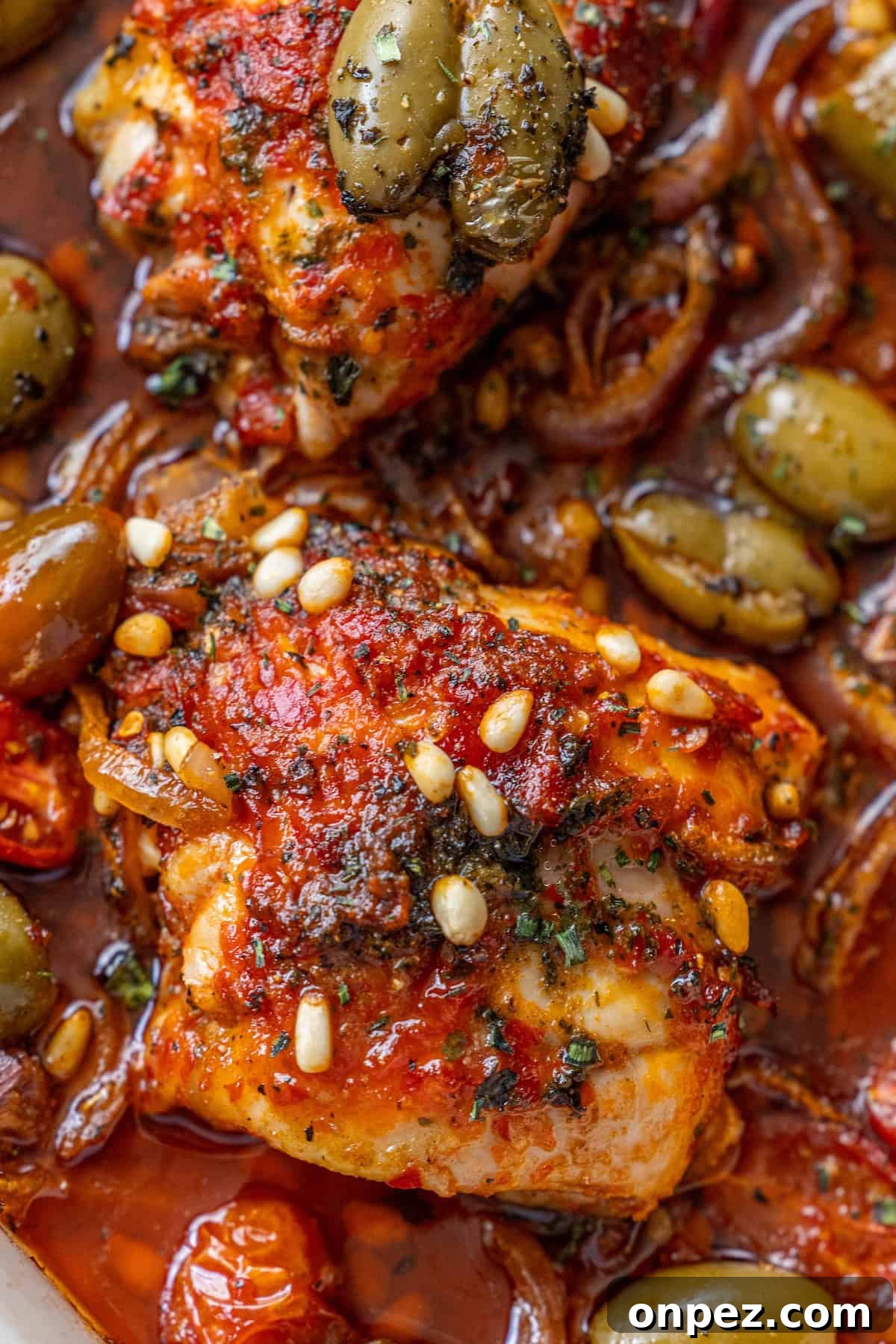 Close-up of cooked chicken topped with tomato sauce, pine nuts, herbs, and green olives in a rich, saucy mixture.