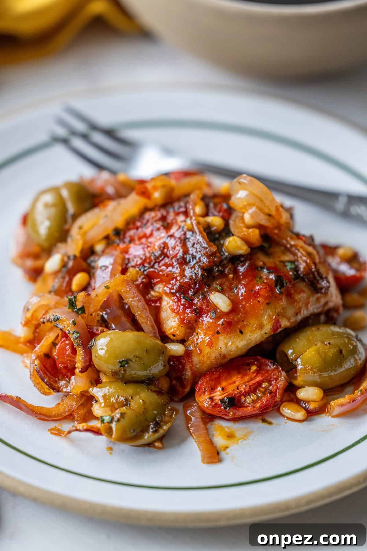 A plate of baked chicken topped with tomato sauce, olives, onions, pine nuts, and herbs, with a fork resting beside the food.