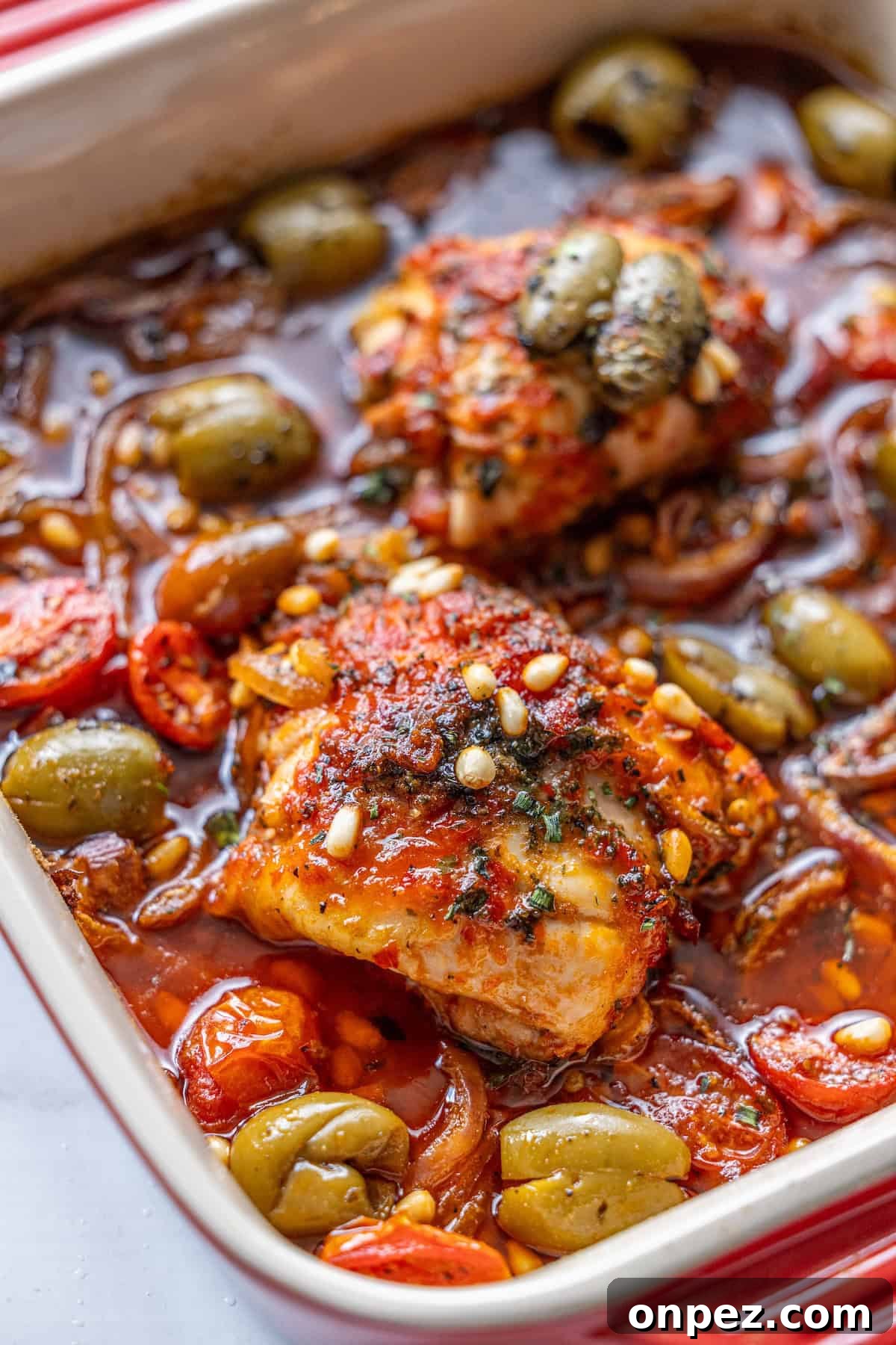 Baked chicken thighs in tomato sauce with green olives, pine nuts, and herbs in a casserole dish.