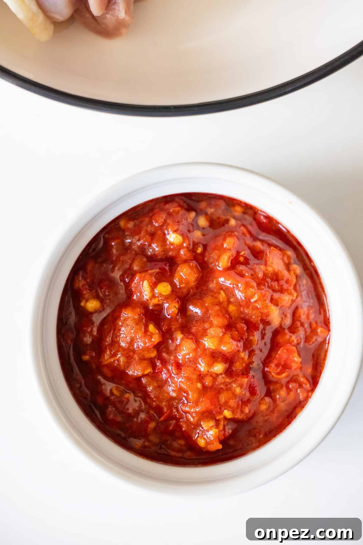 A white bowl filled with chunky red chili paste, perfect for serving alongside spicy chicken thighs, is placed on a white surface.