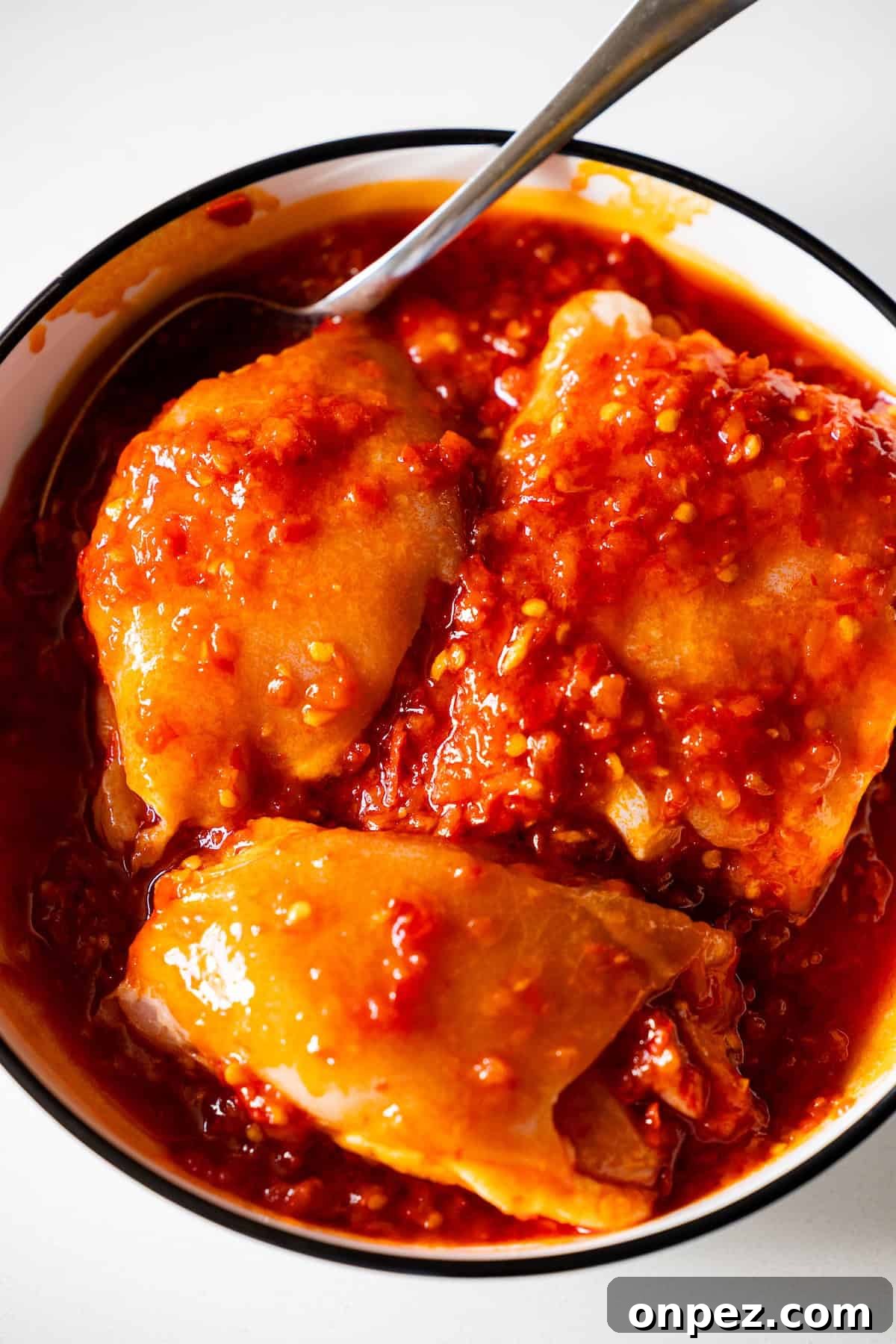 A bowl with three pieces of spicy chicken covered in a thick, red chili sauce, served with smashed olives and a spoon resting on the side.