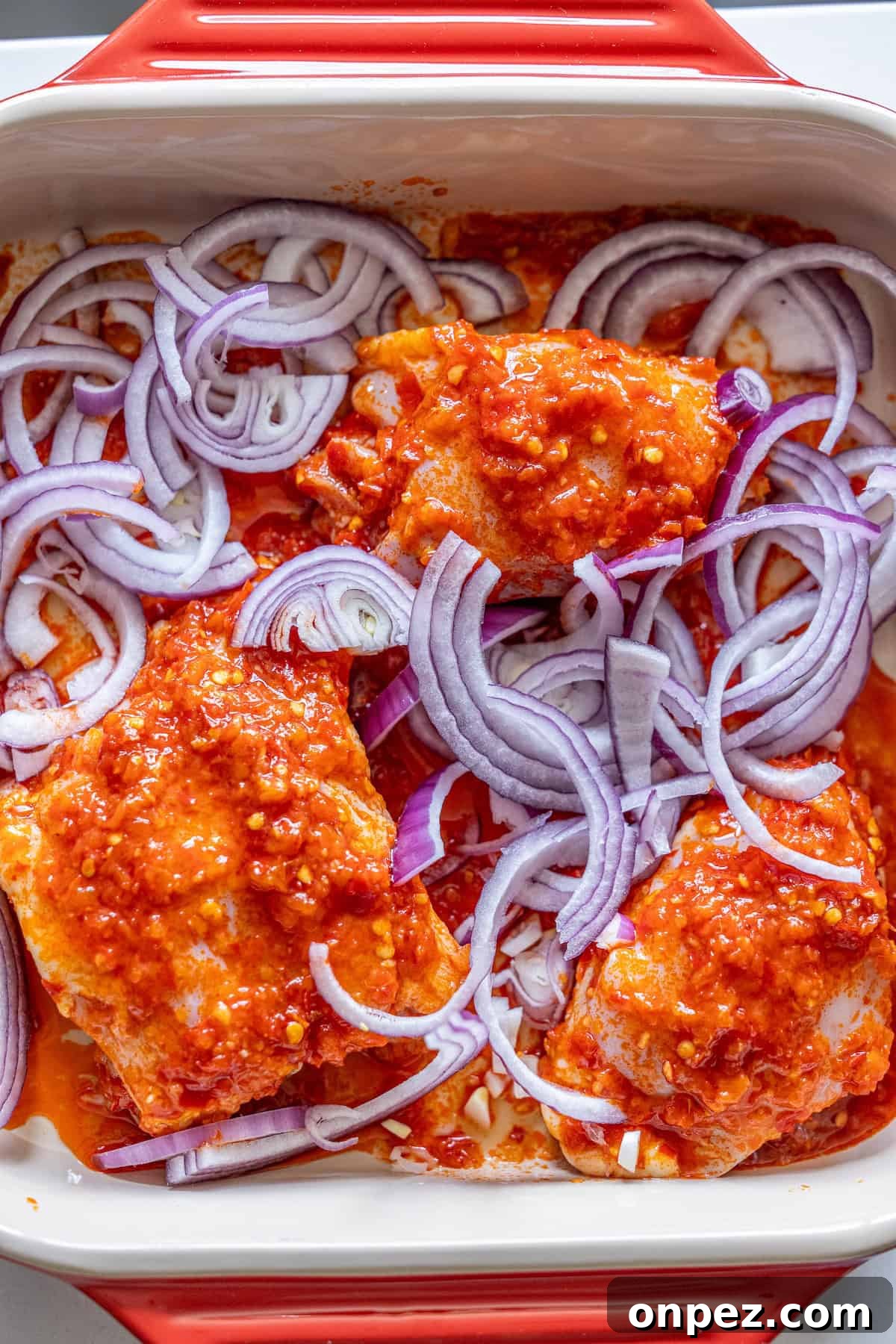 Raw chicken pieces coated in red sauce and topped with sliced red onions in a rectangular baking dish—perfect for making flavorful Harissa Chicken Thighs.