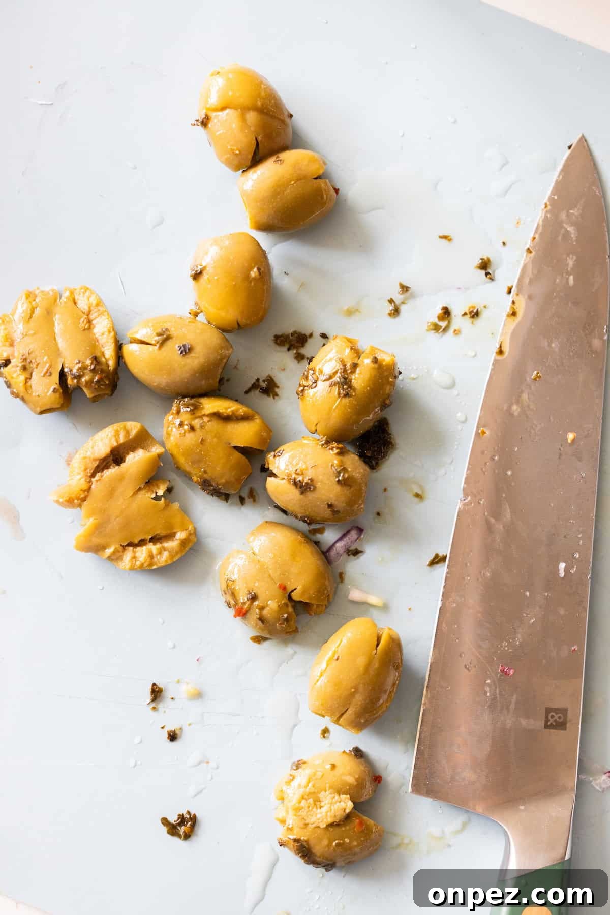 Green olives, some whole and some smashed olives, are scattered on a white cutting board next to a large kitchen knife.