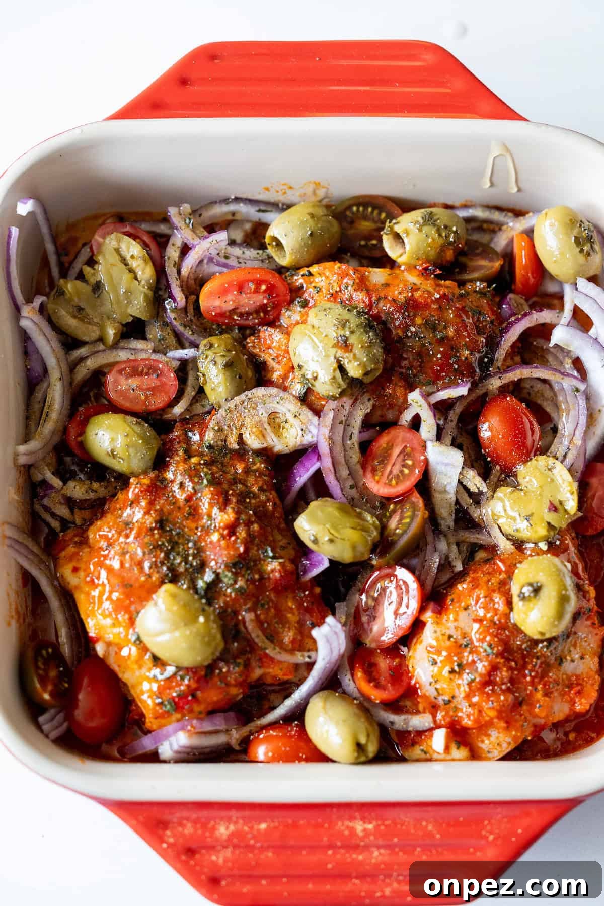 Baking dish with harissa chicken thighs, halved cherry tomatoes, sliced red onions, and smashed olives, ready to be cooked.