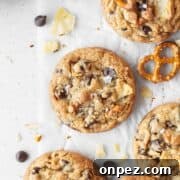 Kitchen sink cookie on a white surface surrounded by more cookies, chocolate chips, crushed potato chips, and pretzels.