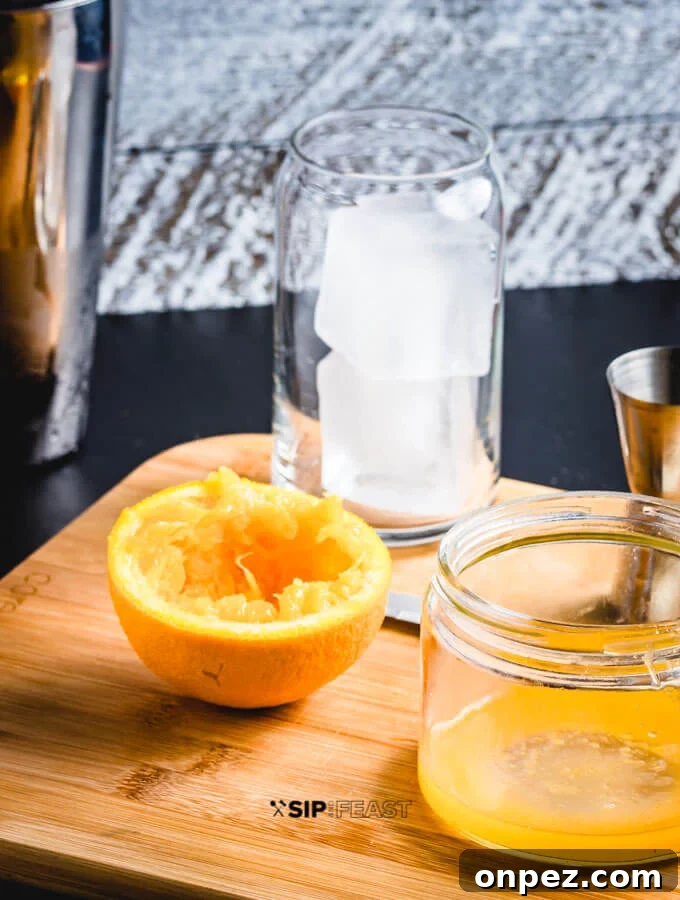 A glass of ice, a cocktail jigger, fresh squeezed orange juice in a citrus juicer and half an orange.