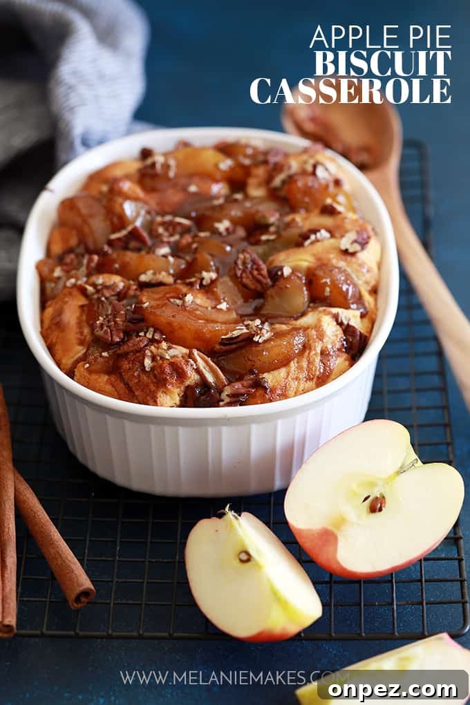 An Apple Pie Biscuit Casserole rests on a wire cooling rack with sliced apples, a wooden spoon and cinnamon sticks.