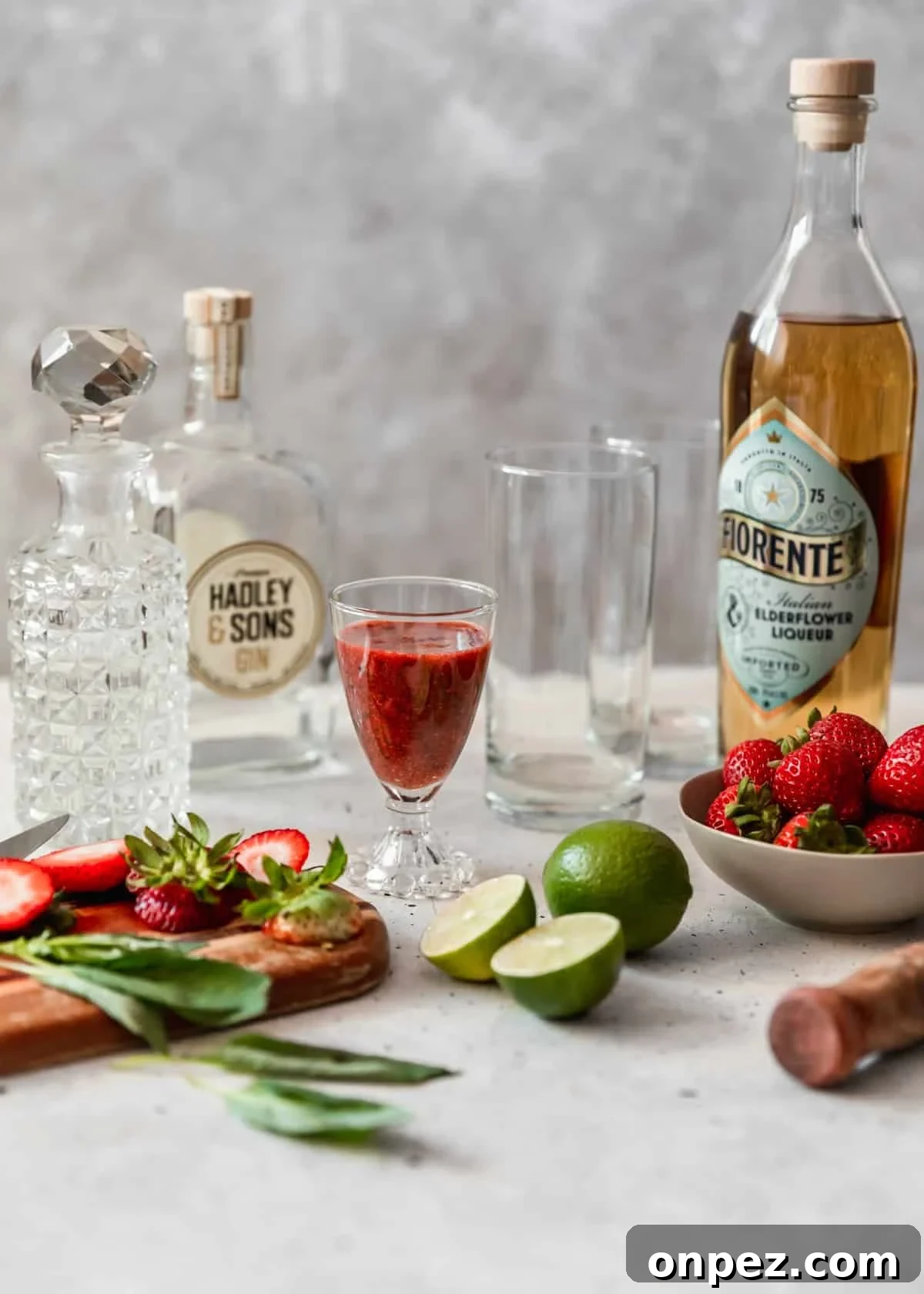 A glass of red juice, bowl of berries, alcohol bottles, a wood muddler, limes, and highball glasses on a counter.