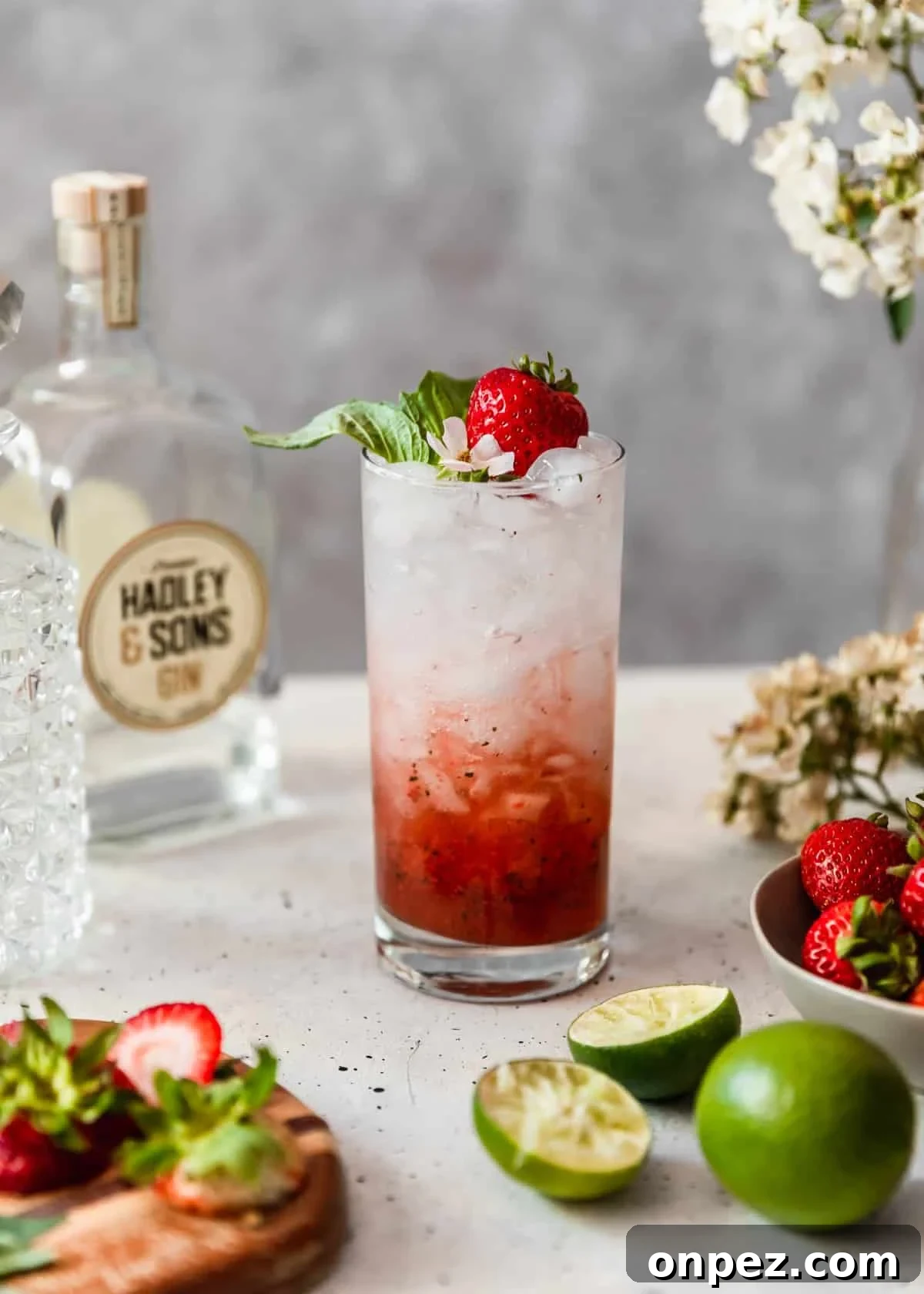 A side image of a strawberry gin smash garnished with a strawberry, basil, and a white flower next to a bowl of strawberries and halved limes.