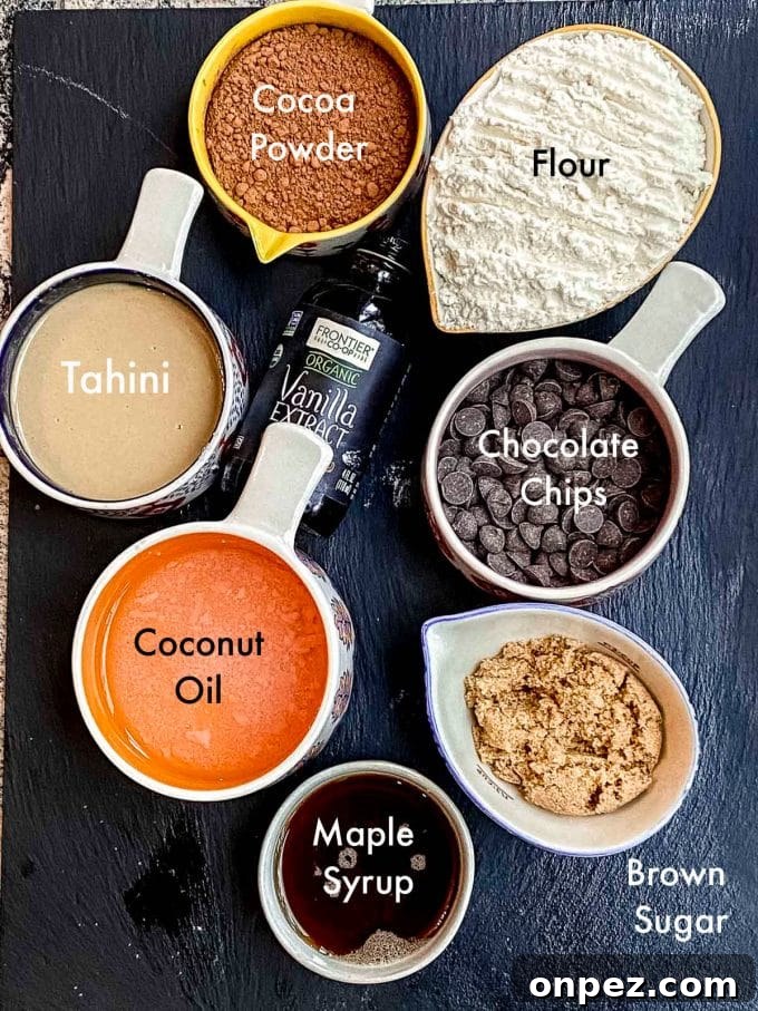 Labeled ingredients to make double chocolate chip cookies: flour, brown sugar, cocoa powder, chocolate chips, vanilla extract, tahini, maple syrup and coconut oil