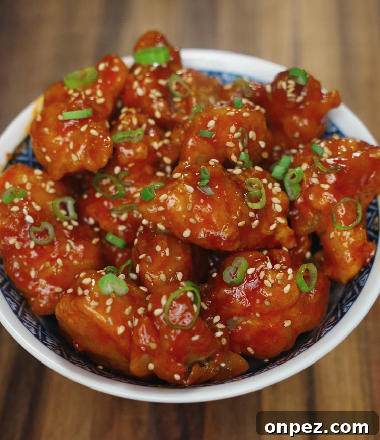 Korean fried chicken coated in a glossy red sauce, garnished with sesame seeds and chopped scallions in a bowl.