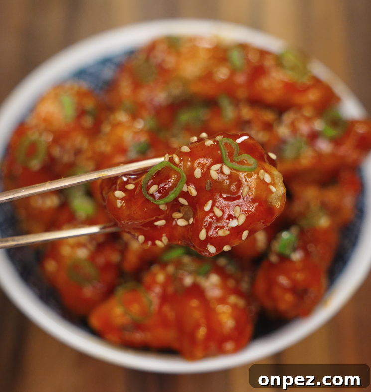 Korean Fried Chicken with Fiery Gochujang Glaze 3 A single piece of spicy Korean fried chicken garnished with sesame seeds and scallions being held with chopsticks.