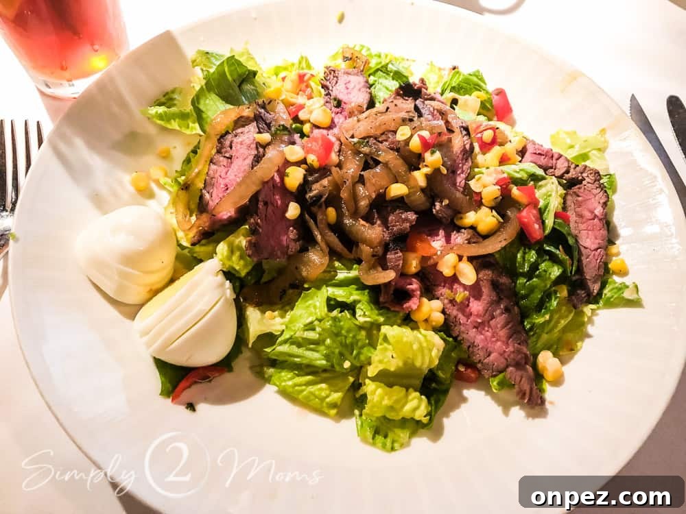 Overhead view of a plate of chopped Romaine lettuce topped with thinly sliced flank steak, pickled onions, corn, diced tomatoes, and a hard boiled egg