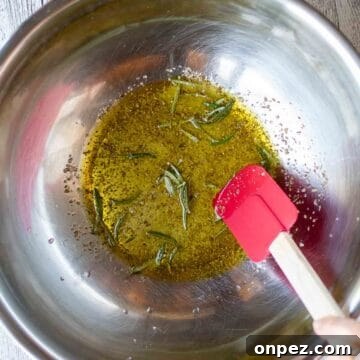 Oil and herbs in a stainless steel bowl with a red spatula viewed from above.