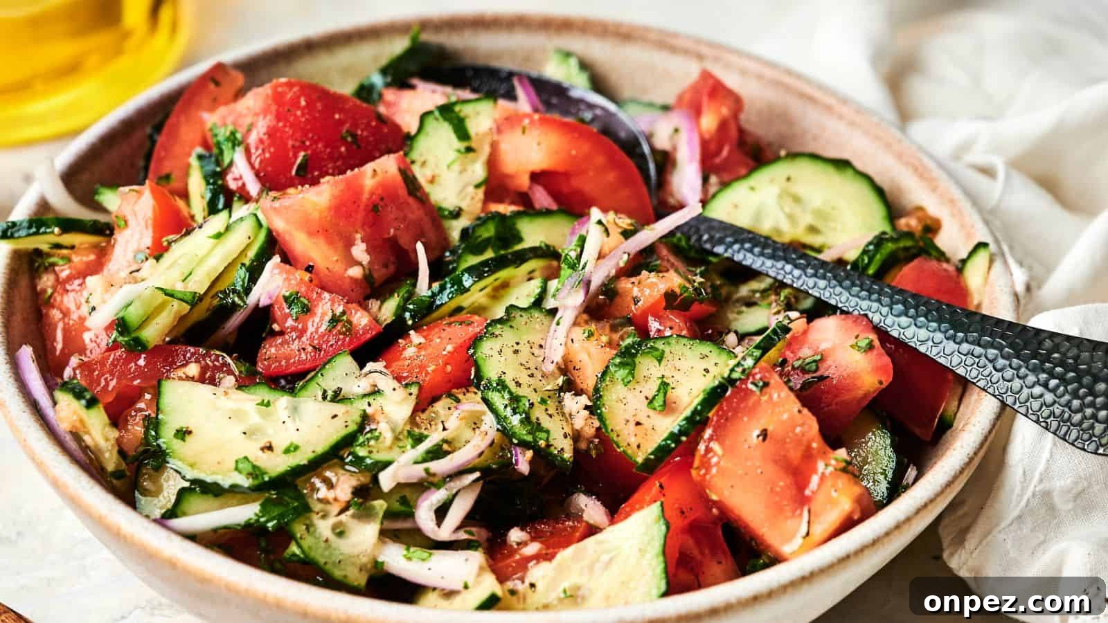 A bowl of fresh salad with sliced tomatoes, cucumbers, red onions, and herbs, seasoned with black pepper, with a serving spoon.