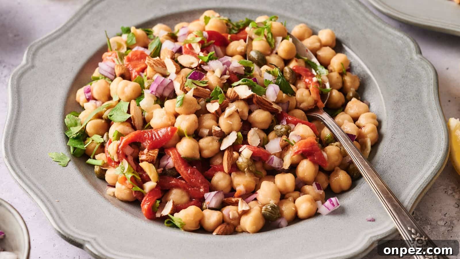 A plate of chickpea salad with chopped red onions, roasted red peppers, herbs, and sliced almonds, served with a spoon.