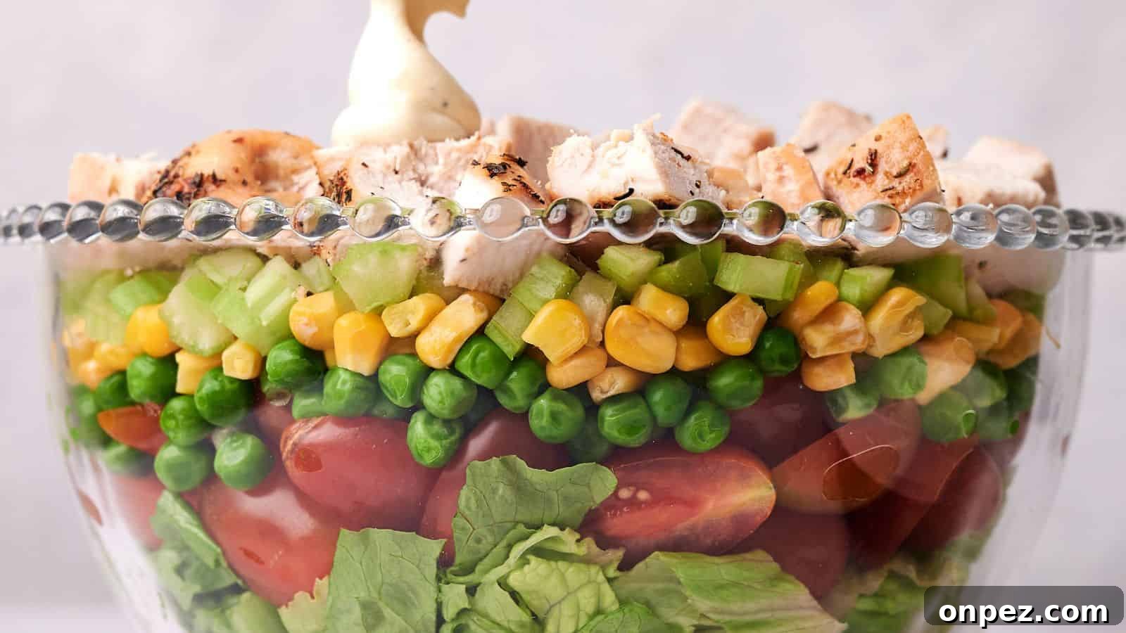 A clear glass bowl layered with lettuce, cherry tomatoes, green peas, corn, diced celery, and topped with chunks of grilled chicken, garnished with a dollop of mayonnaise.