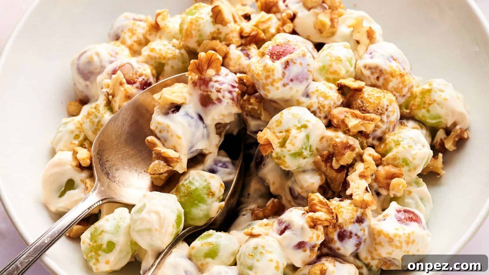 A bowl of grape salad with green and red grapes coated in creamy dressing, topped with chopped walnuts and brown sugar, with a spoon for serving.