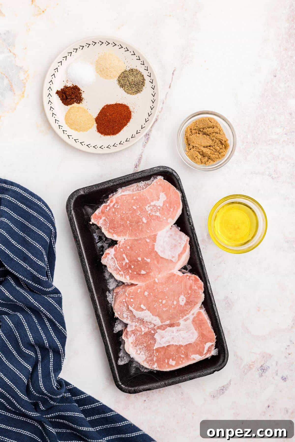 Frozen pork chops on a marble table with olive oil and seasonings for the rub measured out in small dishes.