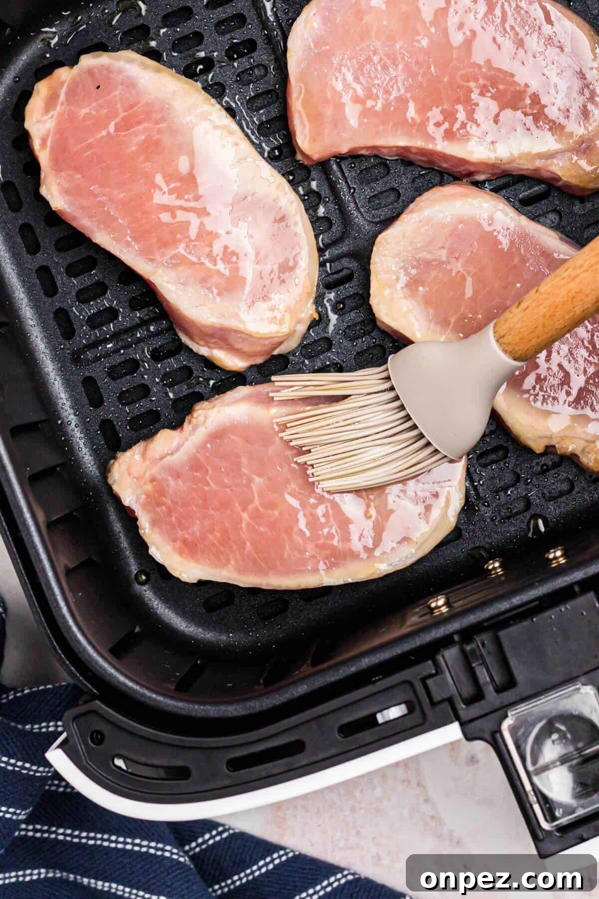 Uncooked pork chops in the air fryer basket being brushed with olive oil before being seasoned.