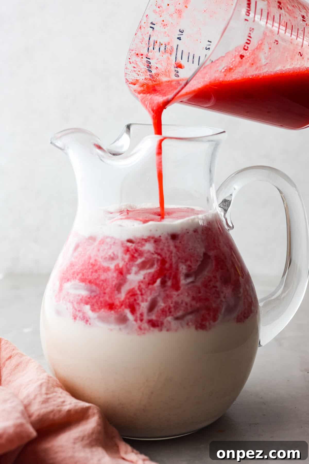 Fresh strawberry puree being poured into creamy horchata. Close view of a large glass pitcher full of horchata with the strawberry puree pouring in.