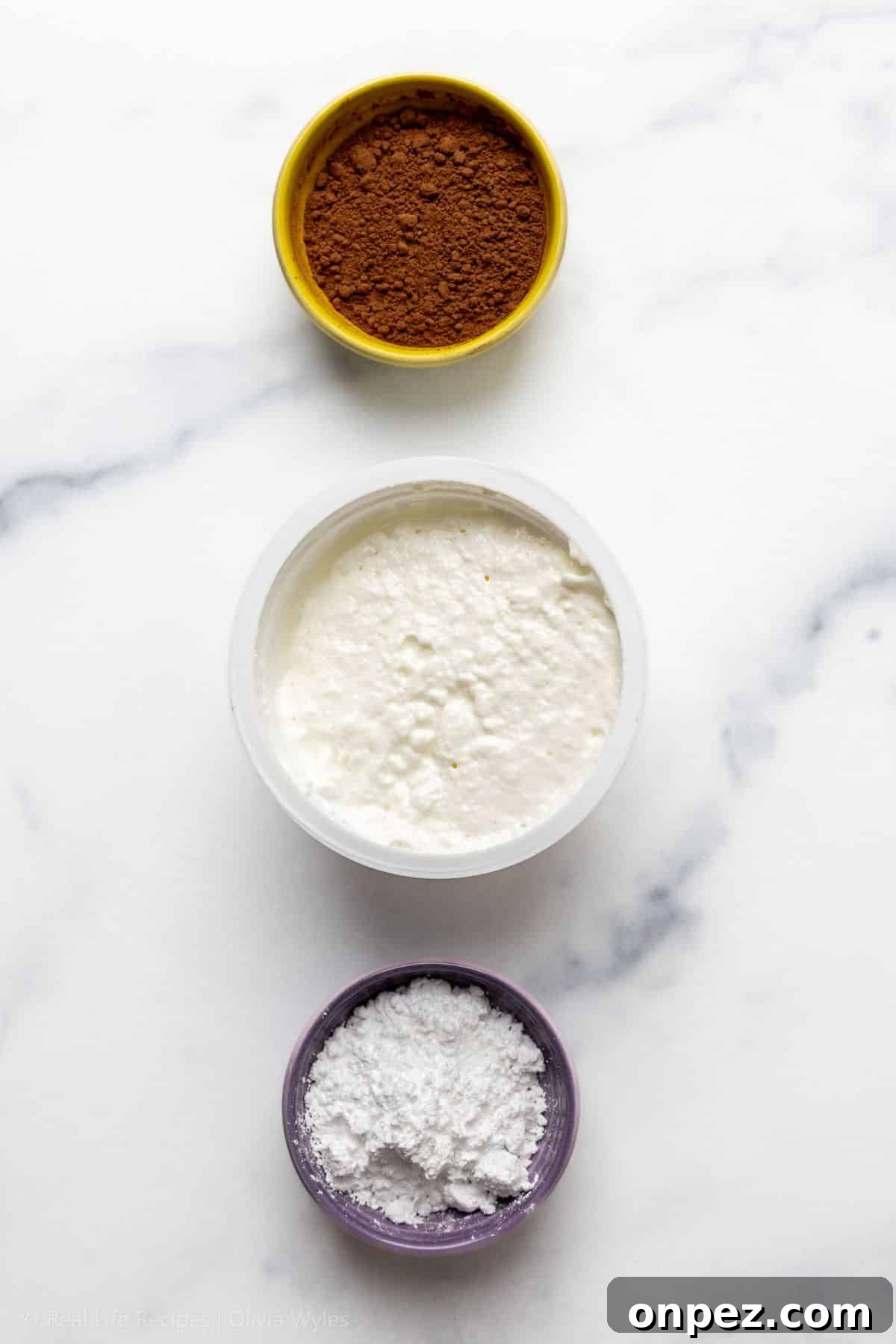 Three bowls of cottage cheese chocolate pudding ingredients on marble: cocoa powder, cottage cheese, and powdered sweetener.