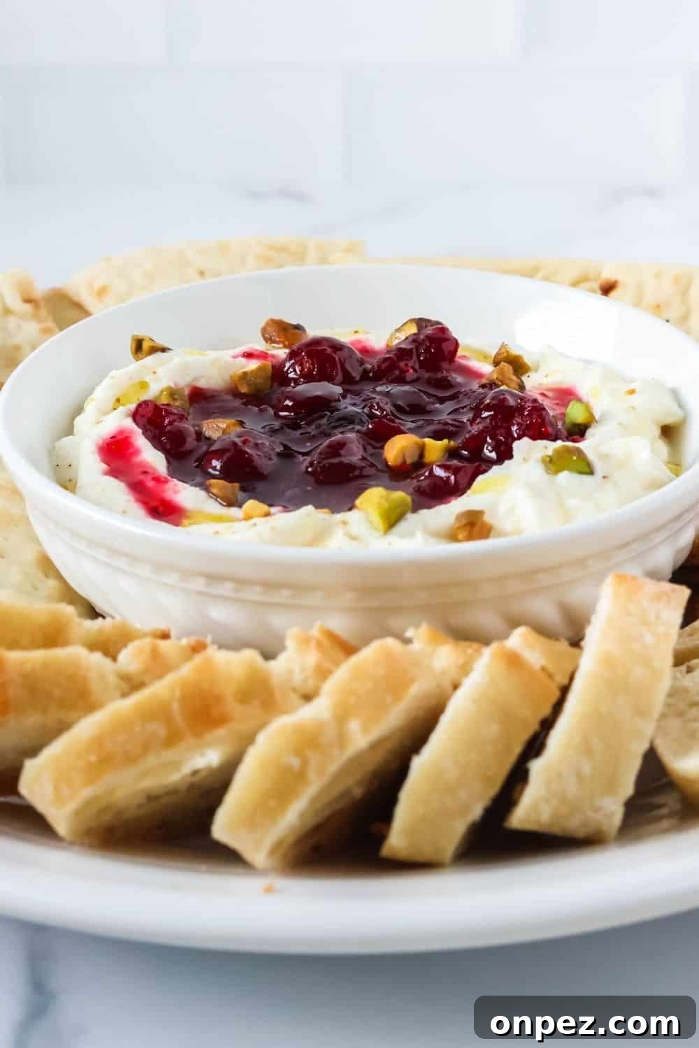 close-up view of cranberry whipped ricotta topped with pistachio nuts. Sliced baguette is next to the dip bowl.