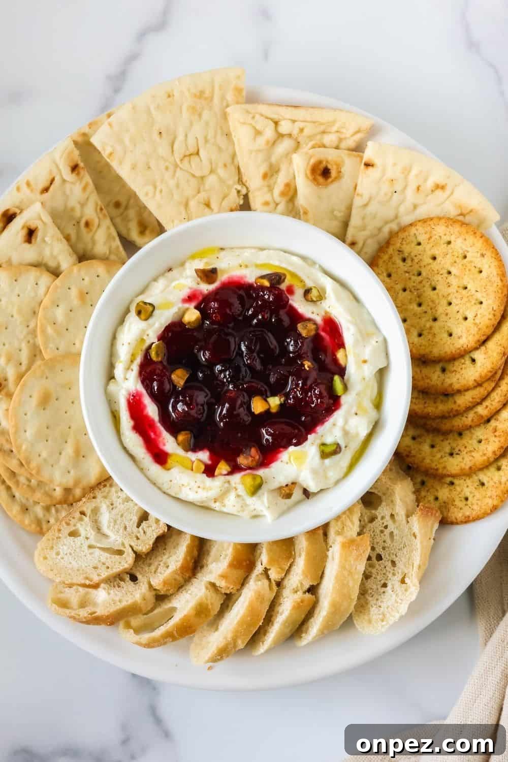 overhead view of a white bowl of cranberry whipped ricotta for the holidays, surrounded by crackers, pita wedges, and baguette slices.