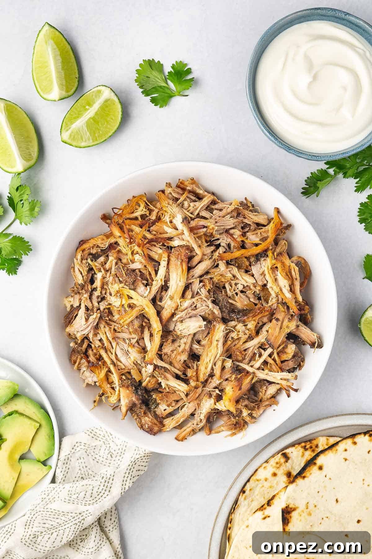 A bowl of shredded slow cooker carnitas with avocados, limes, sour cream, and toasted tortillas on the side.