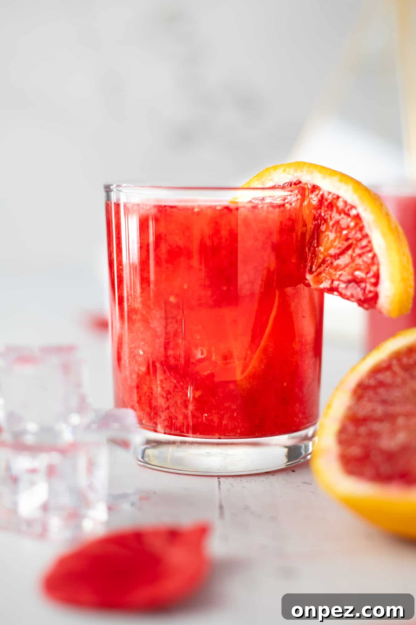 Close up eye level view of a glass of blood orange whiskey sour with ice cubes and rose petals in front