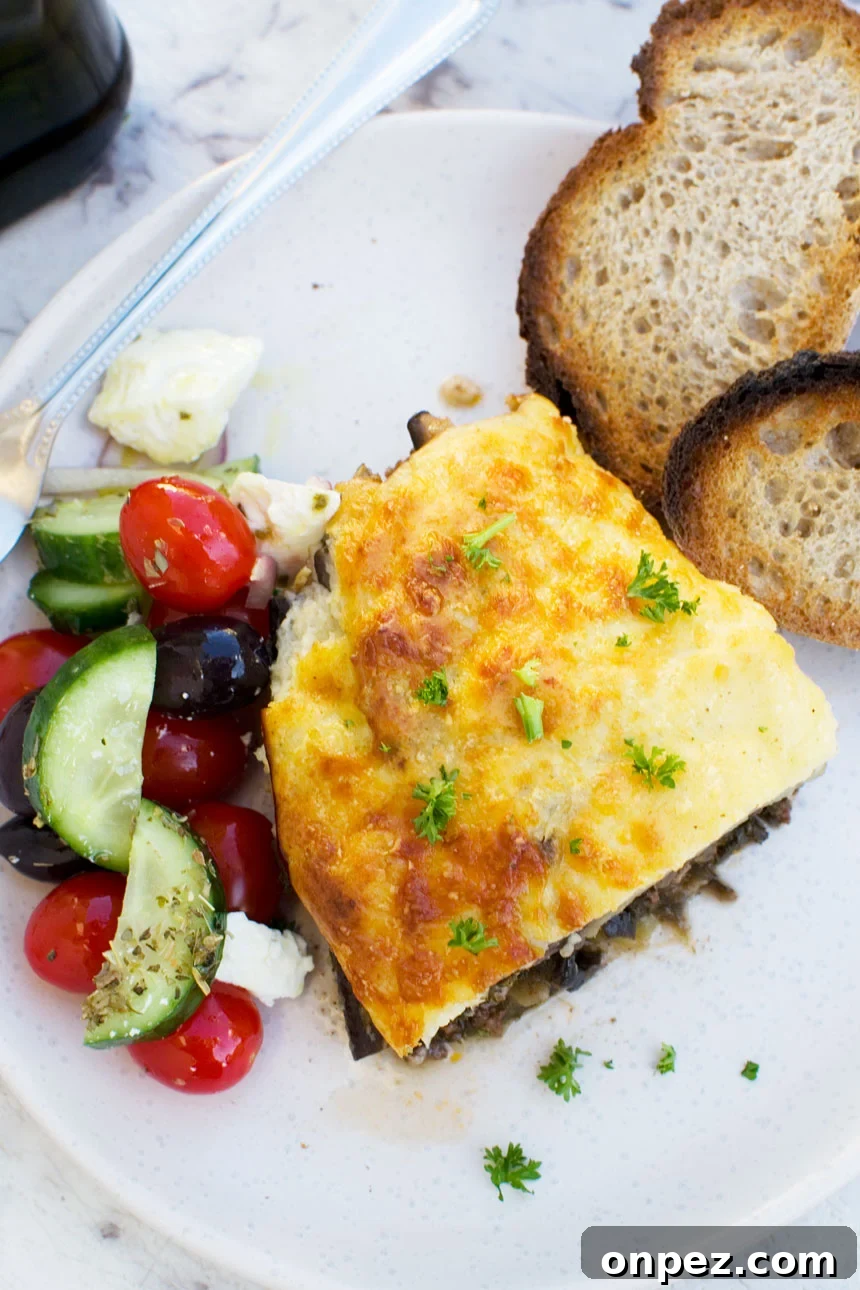 An overhead shot of a piece of easy moussaka on a white plate with bread and Greek salad 