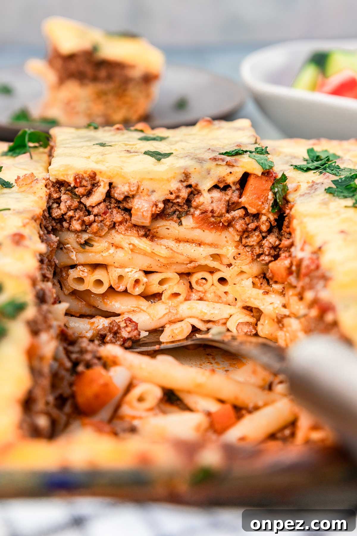 Someone lifting out a square of Greek pastitsio with 3 layers - tube pasta, beef ragu sauce and bechamel sauce, from a large baking dish with a Greek salad and a plate of pastitsio just visible in the background.
