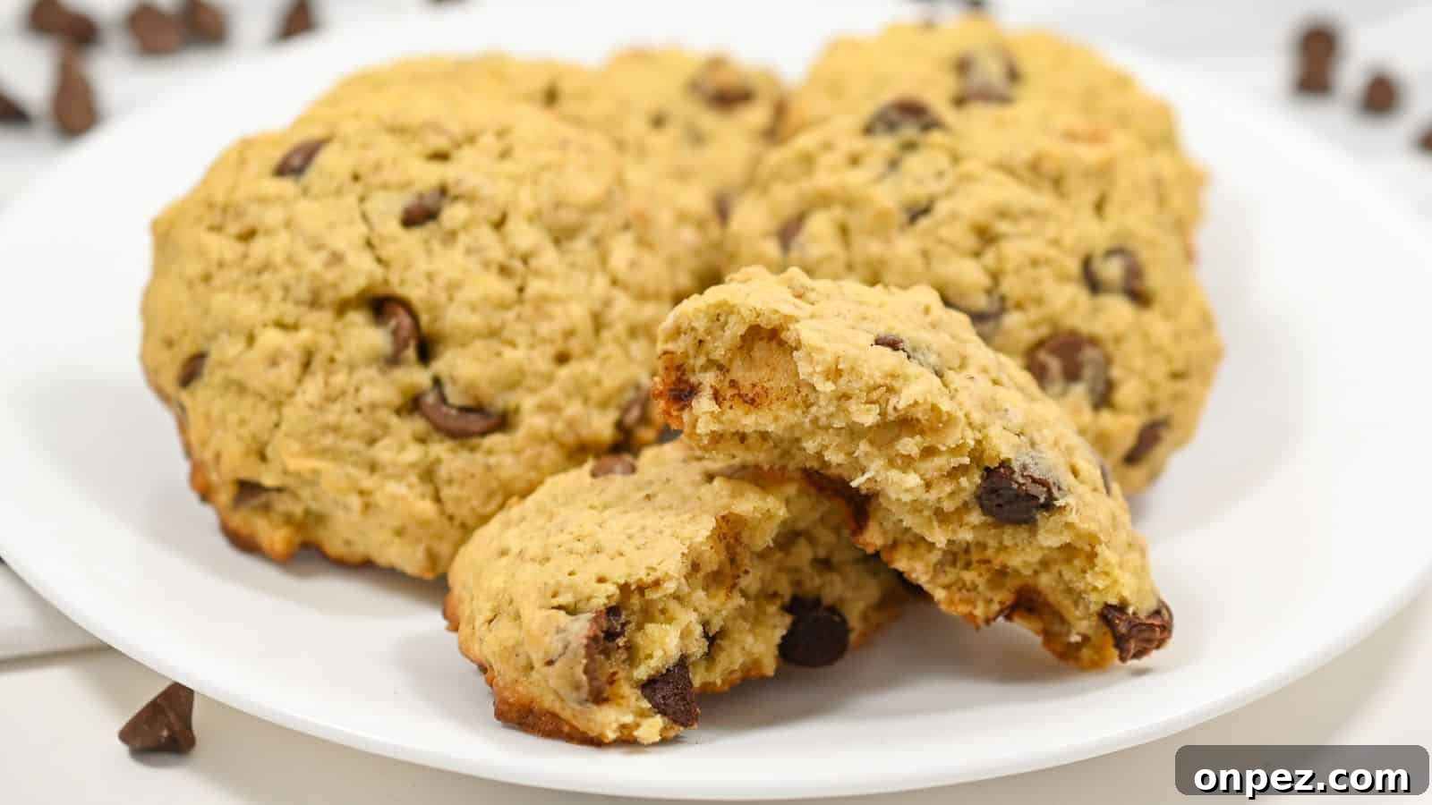 Chocolate oatmeal cookies on a white plate, one broken in half, showing a soft interior, with loose chocolate chips scattered around on a white surface.