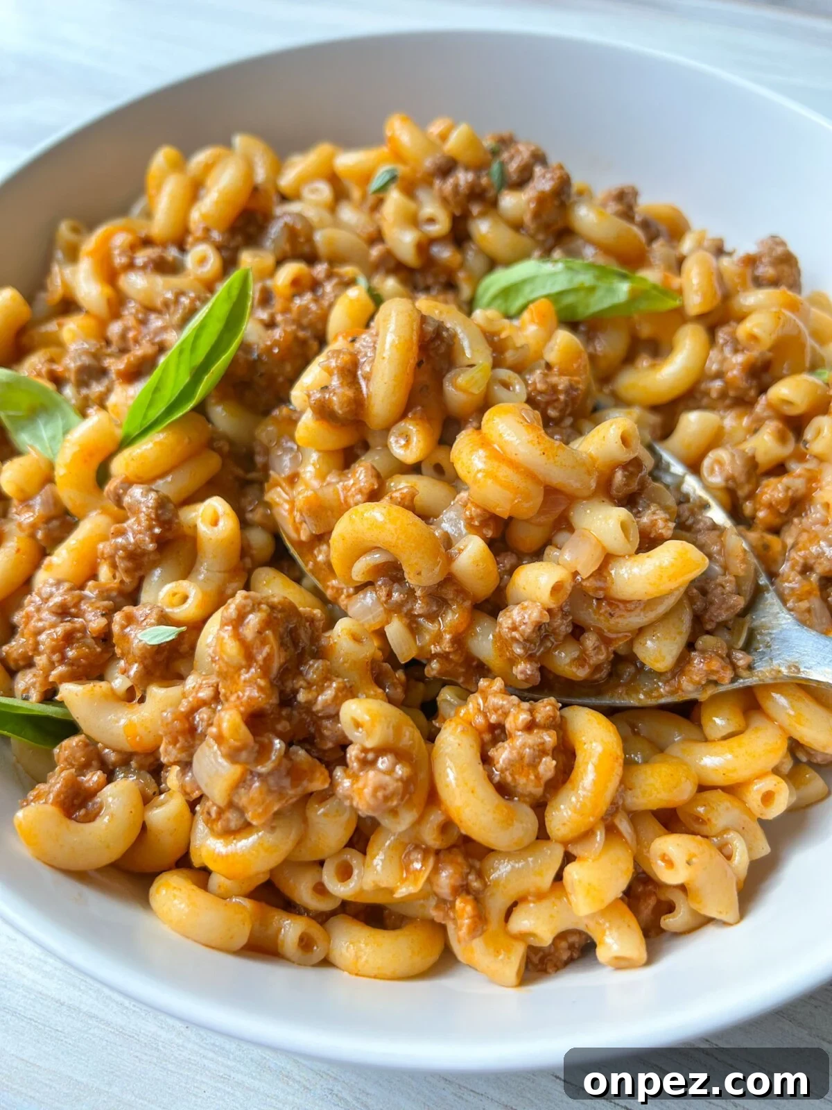 Homemade Hamburger Helper Recipe 2 A white bowl filled with cooked elbow macaroni mixed with a meaty tomato sauce. A few fresh basil leaves garnish the dish. A spoon is partially visible, resting in the macaroni, which looks flavorful and hearty.
