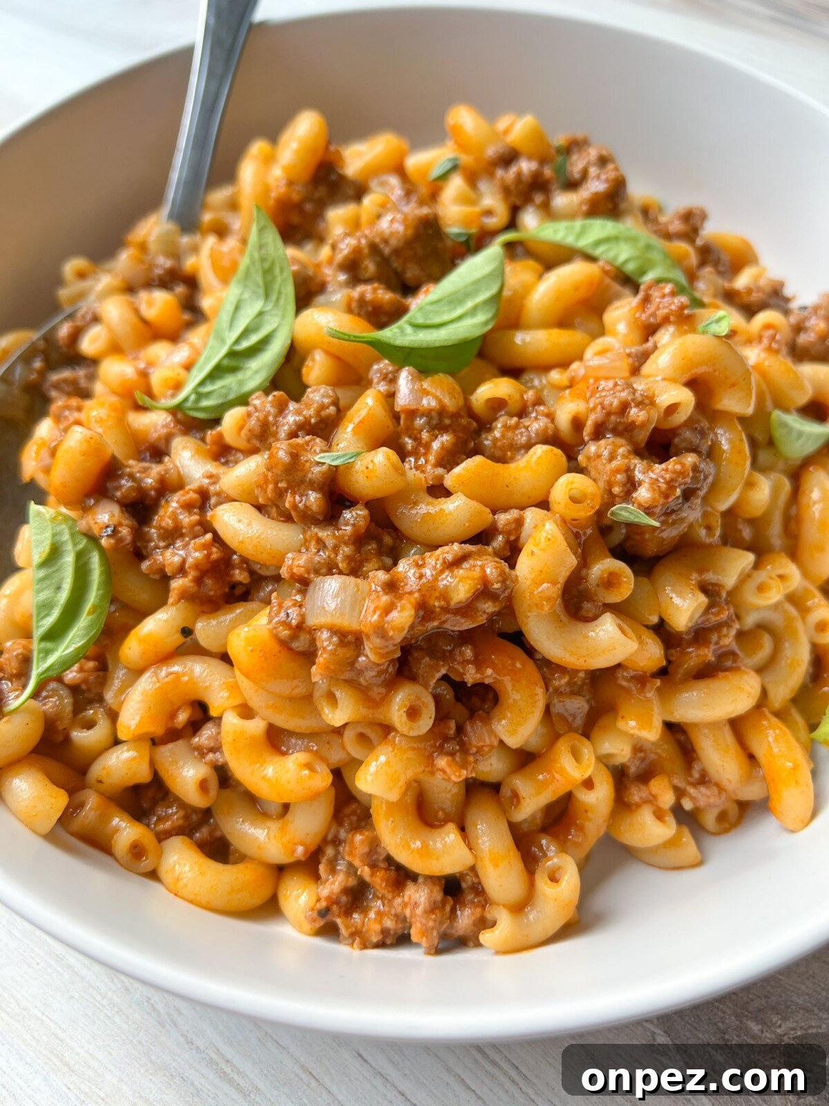 Homemade Hamburger Helper Recipe 3 A white bowl filled with creamy macaroni beef pasta garnished with fresh basil leaves. The pasta is coated in a rich, savory sauce and chunks of ground beef are mixed throughout. A silver fork rests on the side of the bowl.