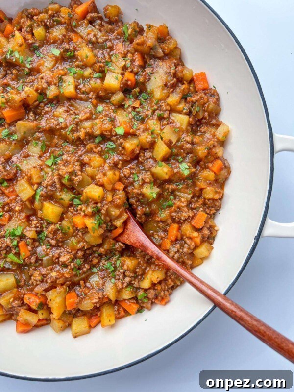 Homemade Hamburger Helper Recipe 5 A close-up of a white pan filled with a hearty ground beef stew, reminiscent of Moussaka with Potatoes, featuring diced potatoes, carrots, and herbs, with a wooden spoon resting inside the mixture.