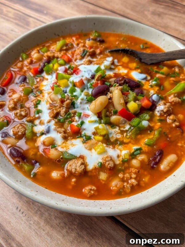 Homemade Hamburger Helper Recipe 6 A bowl of hearty chili with ground meat, red and white beans, chopped peppers, green onions, and a swirl of sour cream on top, served with a wooden spoon on a wooden table.