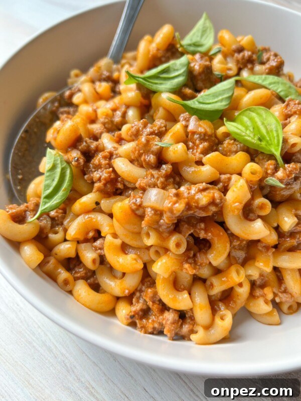 Homemade Hamburger Helper Recipe 9 A close-up of macaroni pasta mixed with ground meat in a tomato-based sauce, reminiscent of classic Hamburger Helper, garnished with fresh basil leaves and served in a white bowl with a spoon.