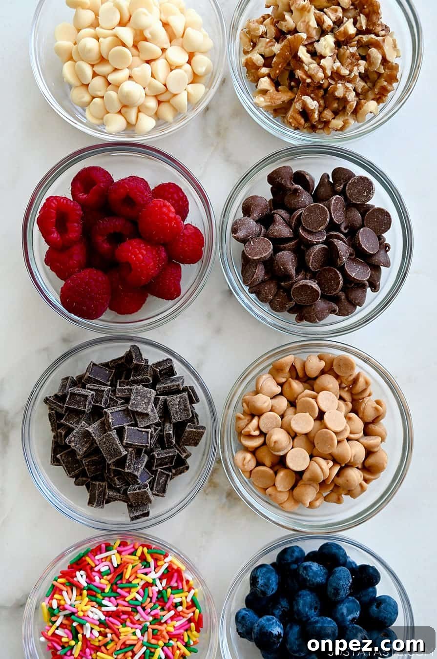 A top-down view of small bowls containing white chocolate chips, walnut pieces, raspberries, chocolate chips, chocolate chunks, peanut butter chips, rainbow sprinkles and blueberries