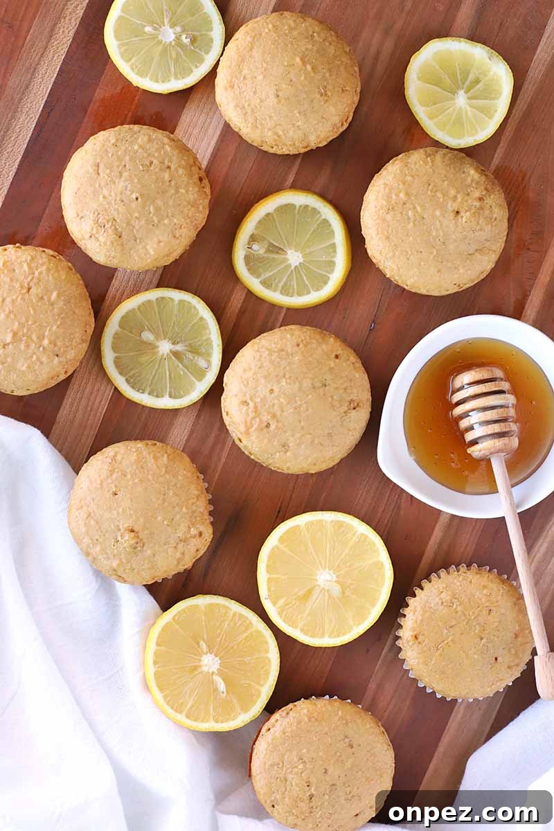 Vertical overhead image of muffins, lemon slices, a small ceramic dish of honey with a wooden dipper, and a white cloth on a brown wood surface.
