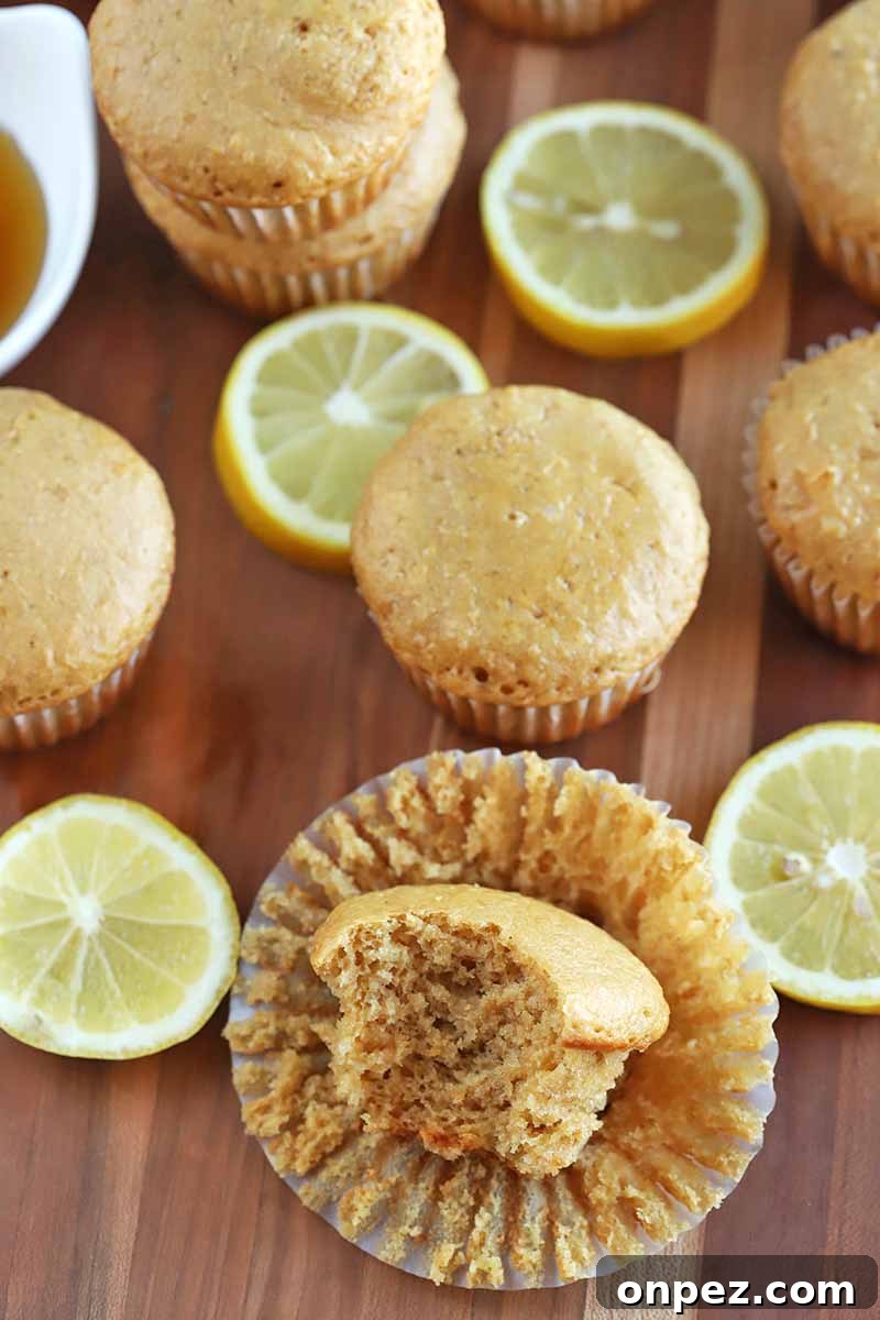 Oblique overhead horizontal image of muffins and lemon slices on a wood surface, with the one in the foreground resting on its paper wrapper with a piece missing to expose the crumb, and a small white dish of honey at the top left of the frame.