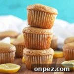 Horizontal image of a stack of three muffins surrounded by several more, with scattered lemon slices on a brown surface, with a white cloth in the background, against a sky blue backdrop.