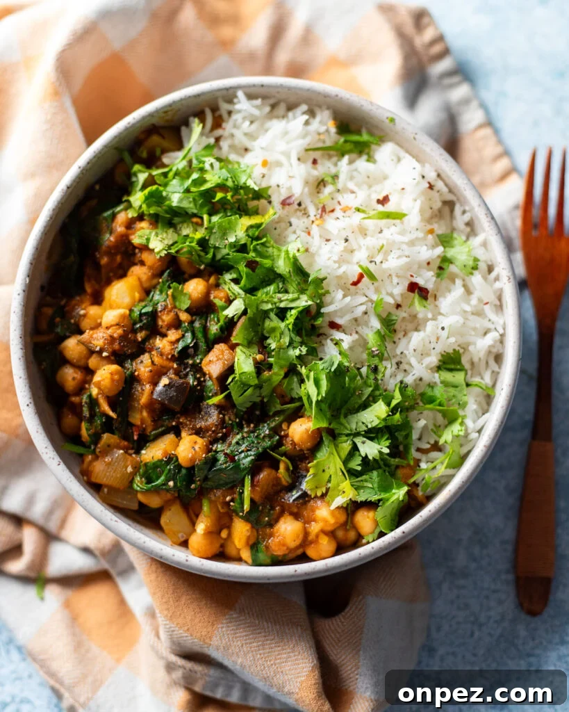 Chickpea, Eggplant and Potato Curry with Spinach Recipe 4 top view of a light blue ceramic bowl half-filled with chickpea, potato, eggplant curry with spinach and aromatics on one side and white rice with green cilantro garnish in the center. The bowl is on a gray tabletop and sitting on a pink and white striped cloth napkin.