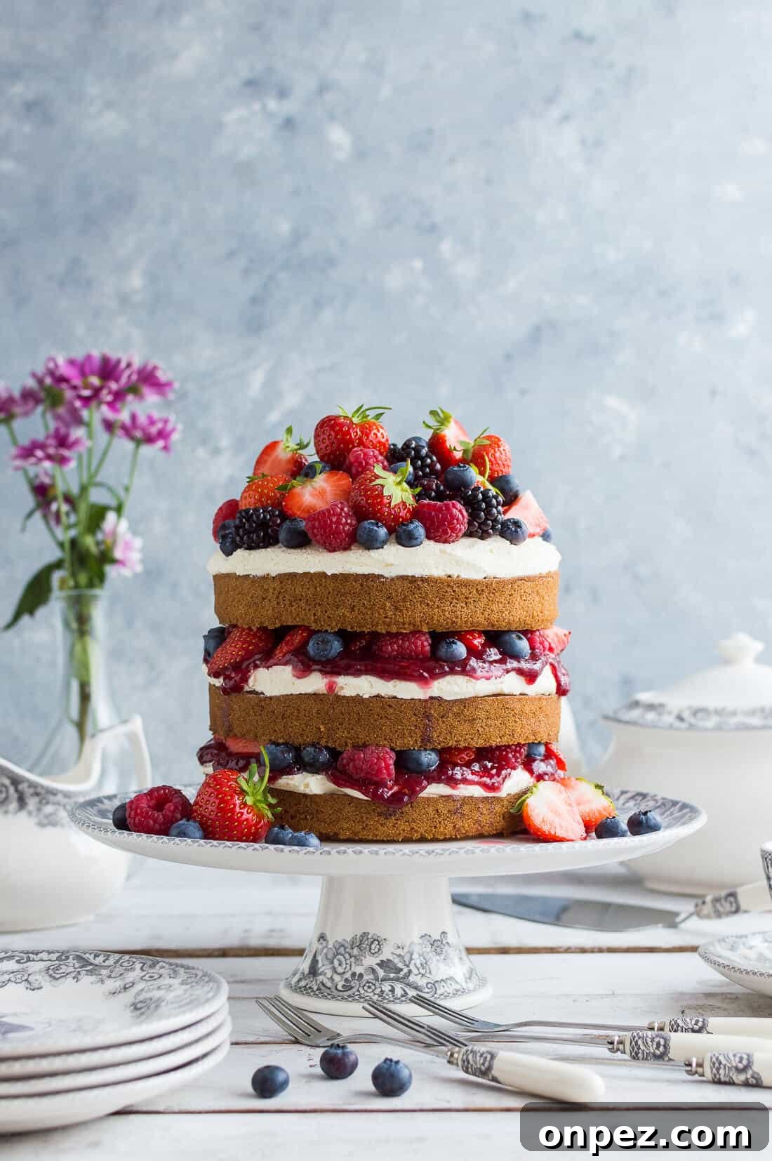 Vegan vanilla cake with berries and jam on a grey and white cake stand with crockery and flowers in the background.